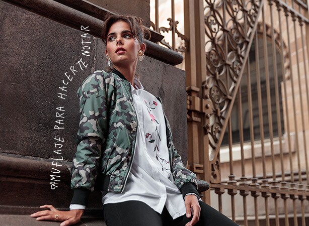 Modelo femenina con chamarra con estampado de camuflaje en colores verde, café oscuro y gris, con contornos negros acanalados en muñecas y cuello y cierre vertical en la parte frontal. Pulsa aquí para comprar tu chamarra, abre en pestaña nueva.