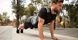 Hombre joven con ropa deportiva Sportline: short negro playera, manga corta y tenis negros, haciendo lagartijas a un lado de un parque con apartado de ejercicios al fondo.